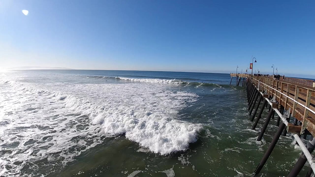 At Pismo Beach, California, foam spreads wider over the dark green water while the pier rail and angled pilings remain on the right, with no visible people.
