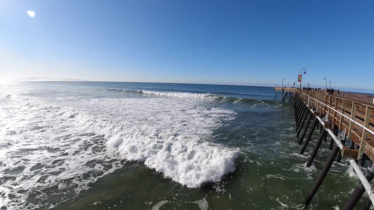 At Pismo Beach, California, a fresh band of whitewater crosses the midground as the pier extends from the right and the clear horizon stays visible, with no visible people.