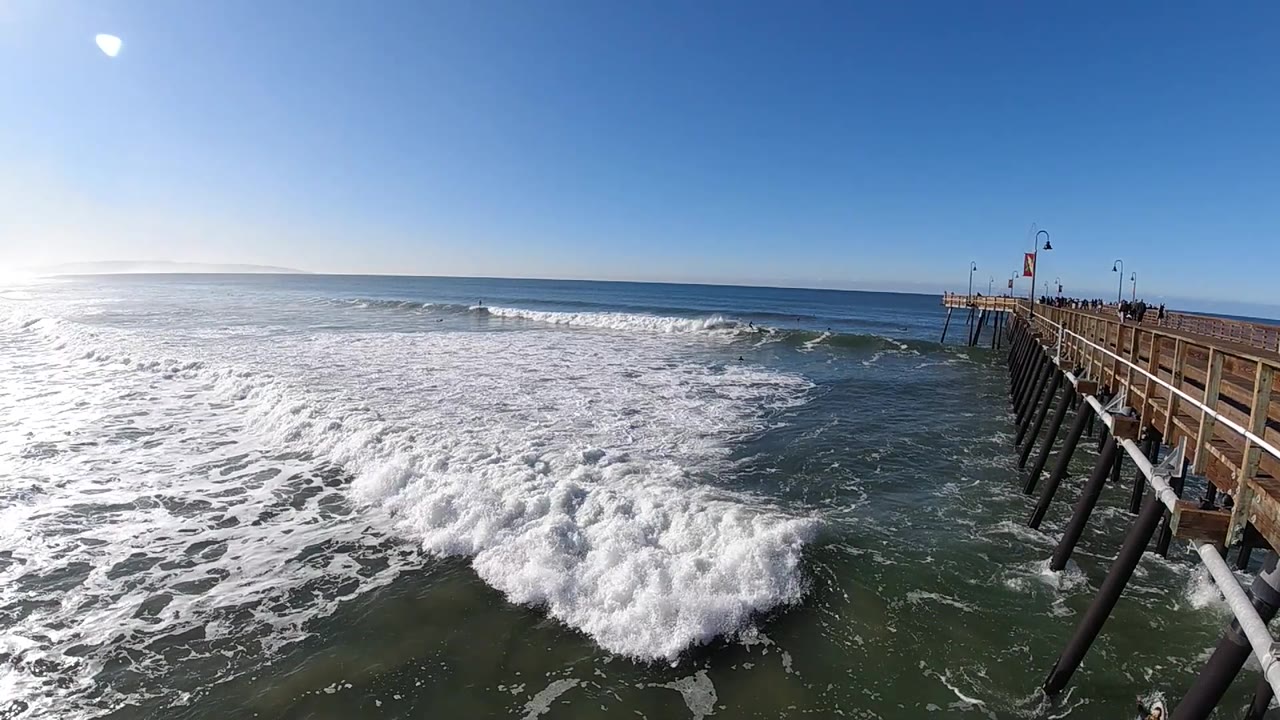At Pismo Beach, California, the whitewater gathers in the lower frame while darker open water appears beside the pier, with no visible people in the sampled view.