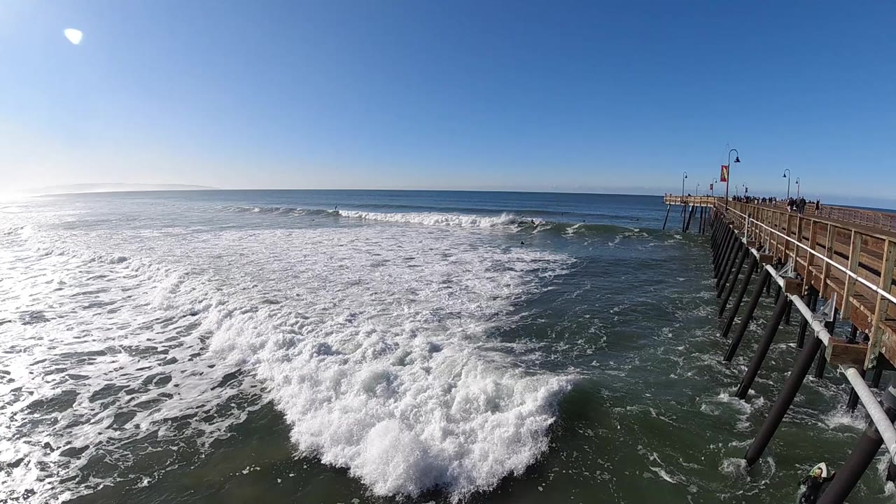 Foamy surf spreads across the foreground beside a wooden pier at Pismo Beach, California, with bright sun flare in the upper left and no discernible people.