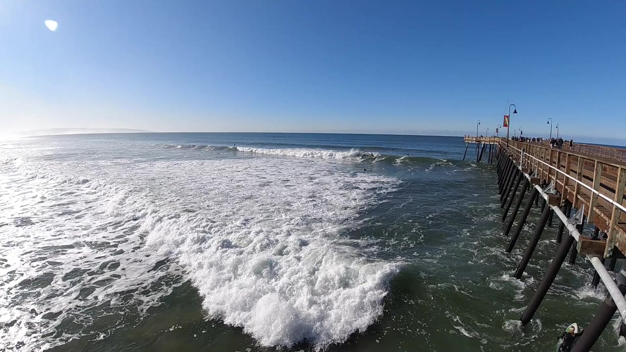A breaking wave pushes whitewater toward the lower frame while the Pismo Beach pier stays along the right edge, with open blue sky above and no discernible people.