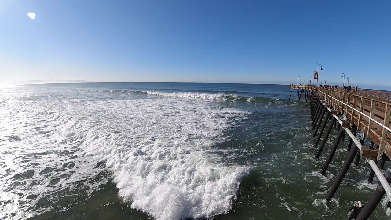 White foam fills more of the foreground at Pismo Beach as the wave wash spreads outward, with the wooden pier receding from the right side and no discernible people.