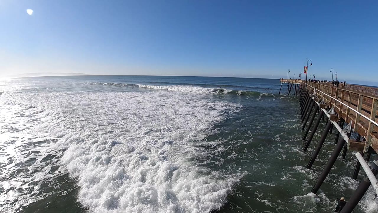 White surf breaking beside a wooden pier under blue sky.