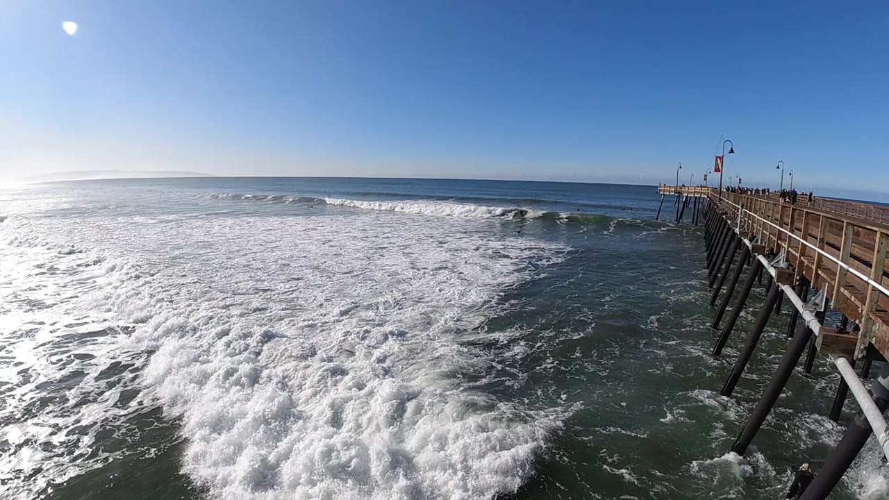 At Pismo Beach, California, the wave line sits farther across the midground while thick white foam fills the lower frame and the pier pilings remain on the right.