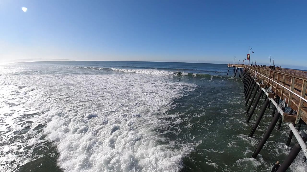 At Pismo Beach, California, broad whitewater covers most of the foreground as open ocean and a low wave line stretch across the horizon beside the pier.