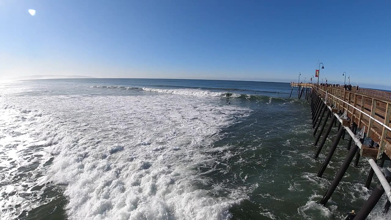 At Pismo Beach, California, foamy surf pushes across the lower left while the wooden pier and dark support pilings angle down the right side of the frame.