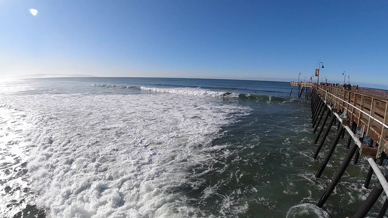 Foam covers much of the foreground as a darker wave breaks near the right-side pier in Pismo Beach, California, with no visible people.