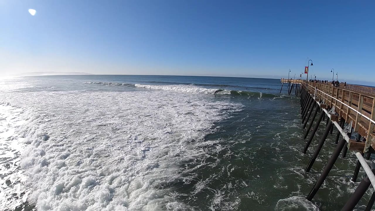 A broad band of white surf pushes across green ocean water beside the pier in Pismo Beach, California, with clear sky and no visible people.