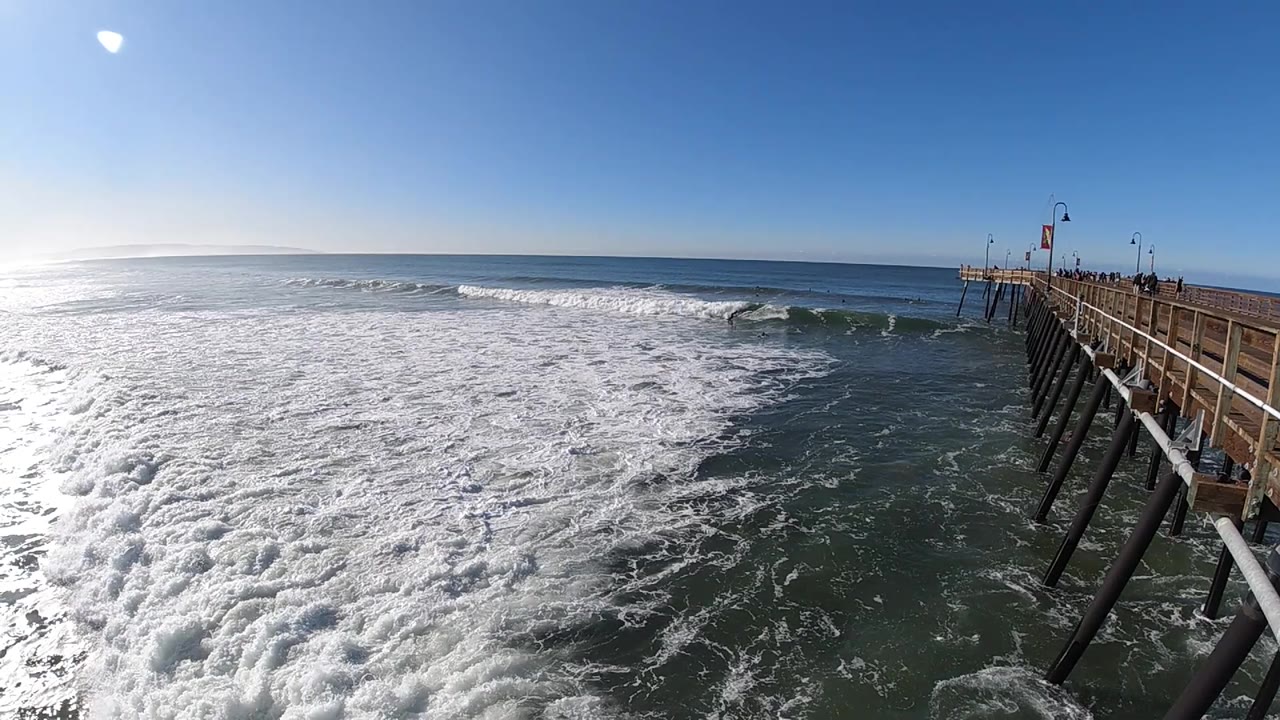 The foreground is packed with bright foam while darker water opens near the right-side pier in Pismo Beach, California, with no visible people.