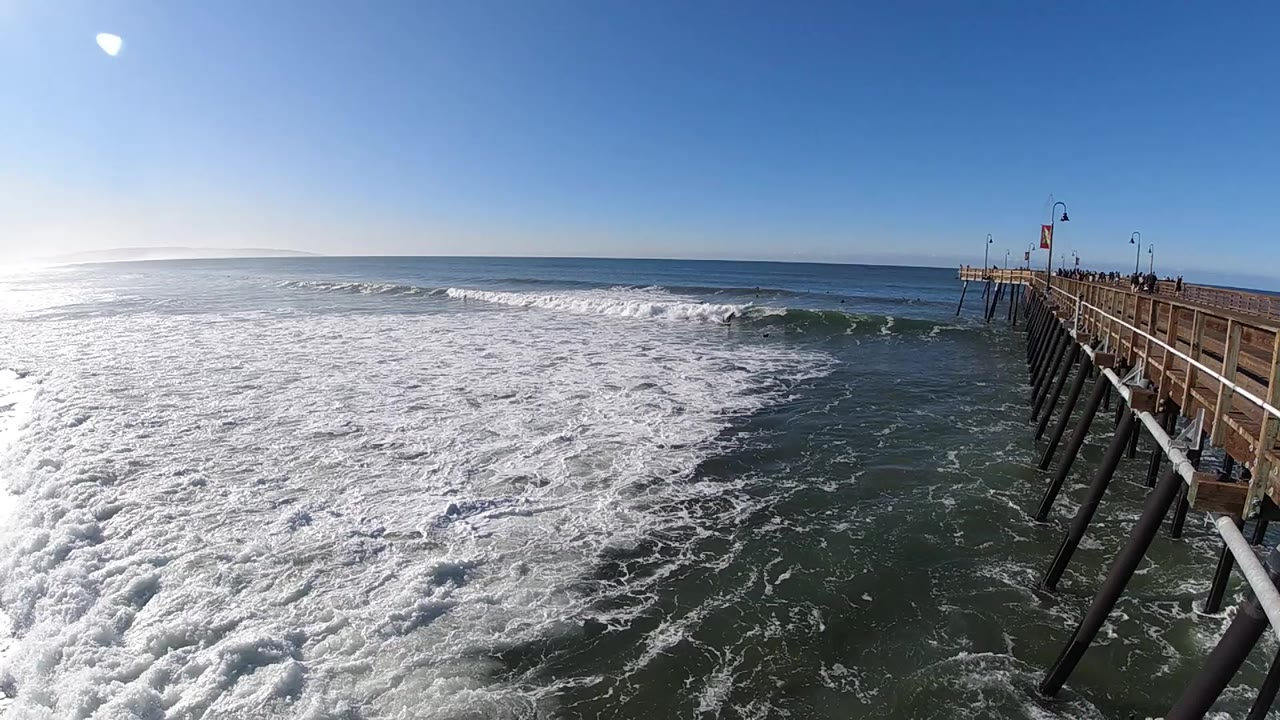 At 0:01 in Pismo Beach, the same wide pier view shows a darker band of water near the pilings and a line of small breaking waves beyond the foam, with no visible people.