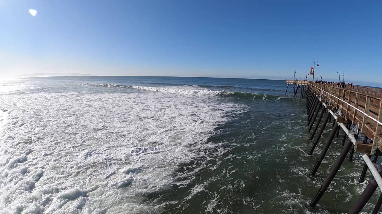 At 0:02 in Pismo Beach, bright whitewater fills the left foreground while the pier recedes from the right edge toward the horizon, with no visible people.