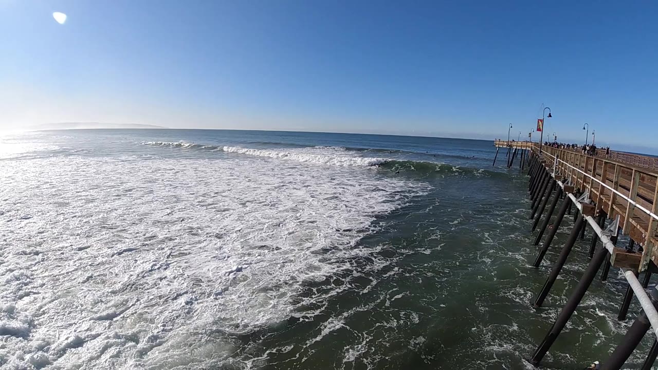 Wide view of foamy Pacific surf at Pismo Beach, California, with a wooden pier along the right edge and small people visible on the rail.