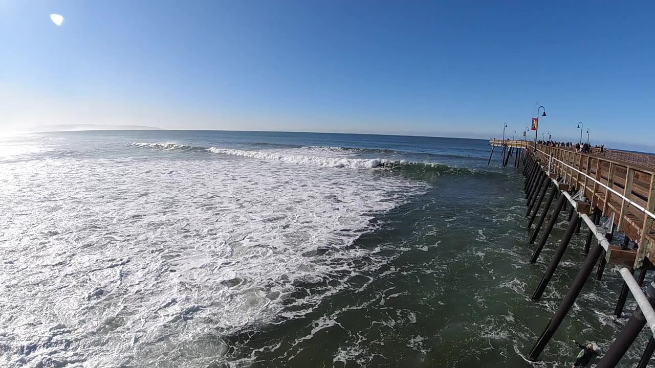 Pismo Beach surf spreads across the foreground beside a wooden pier on the right, with bright ocean glare and no discernible people in view.