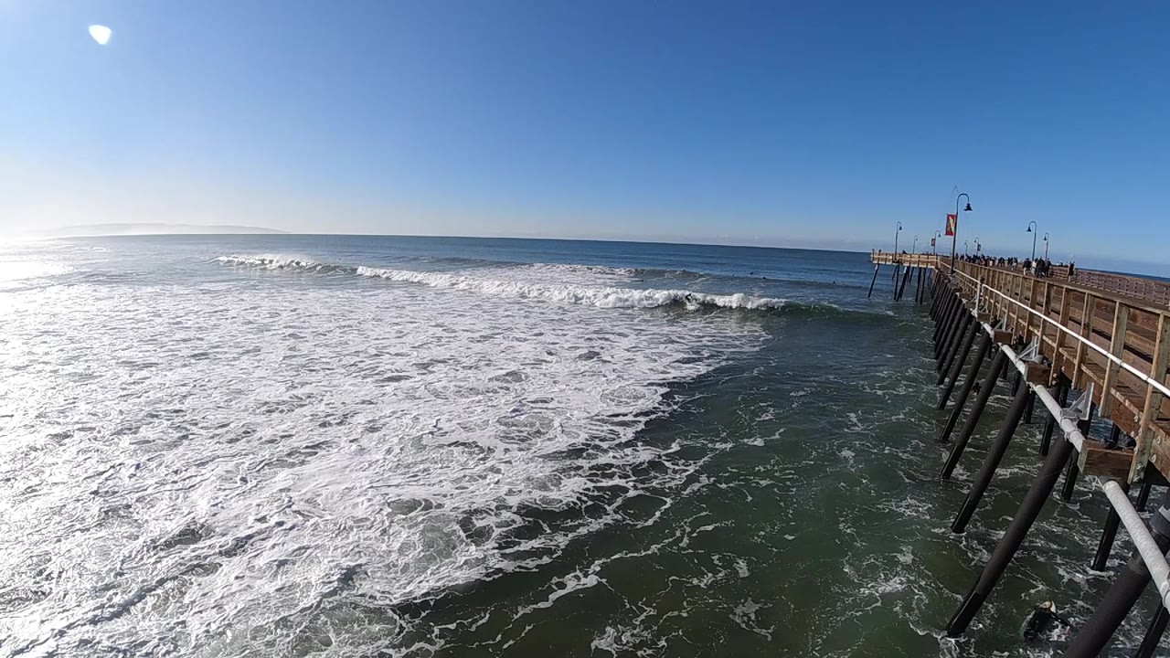 At 0:01 in Pismo Beach, California, the surf line sits slightly farther across the midground, with bright glare on the left water and the pier still framing the right edge.