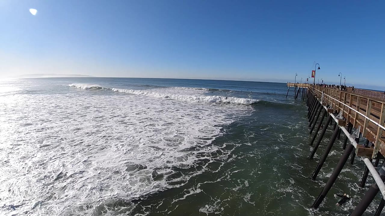 A broad sheet of whitewater spreads across the left and center of the Pismo Beach ocean view while the wooden pier and its pilings remain tight along the right edge, with no visible people.