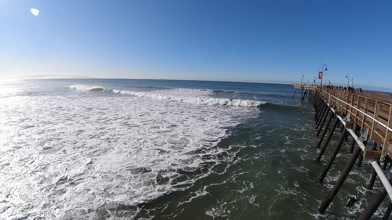 A new band of whitewater rolls across the center-left of the Pismo Beach frame, with darker water below, the wooden pier on the right, and no visible people.