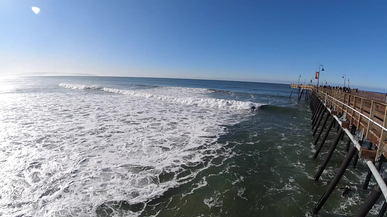 Pismo Beach surf rolls toward the lower left while the pier railings and dark support pilings run along the right side, with no visible people.