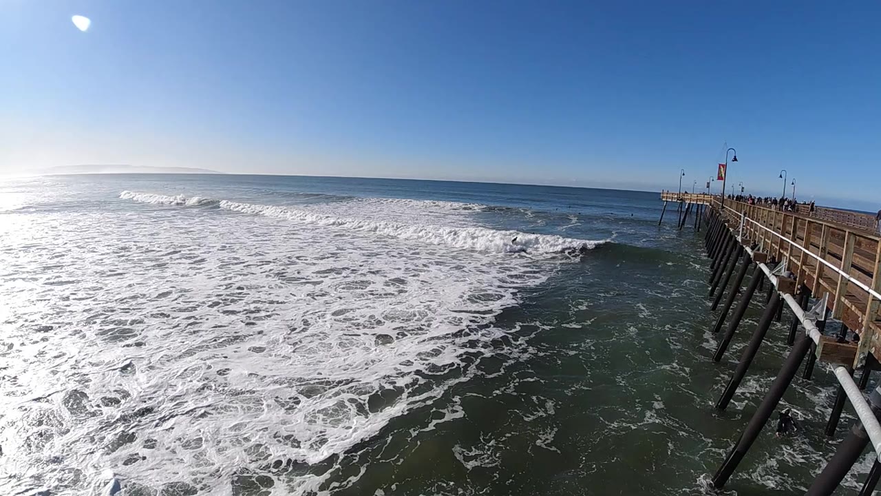 Wide view of foamy surf beside a long wooden pier in Pismo Beach, California, with bright sky, sun glare, and the ocean horizon across the background.