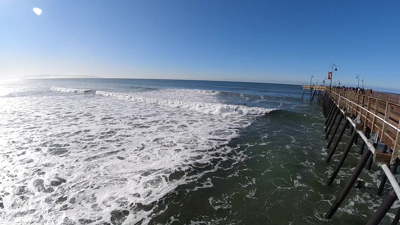 Foamy whitewater spreads across the Pismo Beach shoreline view while the pier remains on the right and the open ocean sits beyond the surf line.