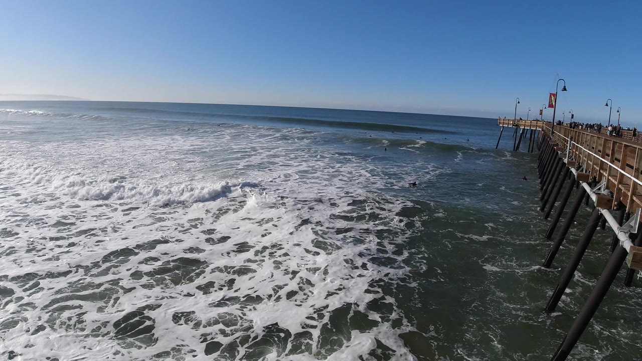 Pismo Beach waves roll through the middle distance as foam covers the lower frame, with the wooden pier and dark pilings along the right edge and no visible people.