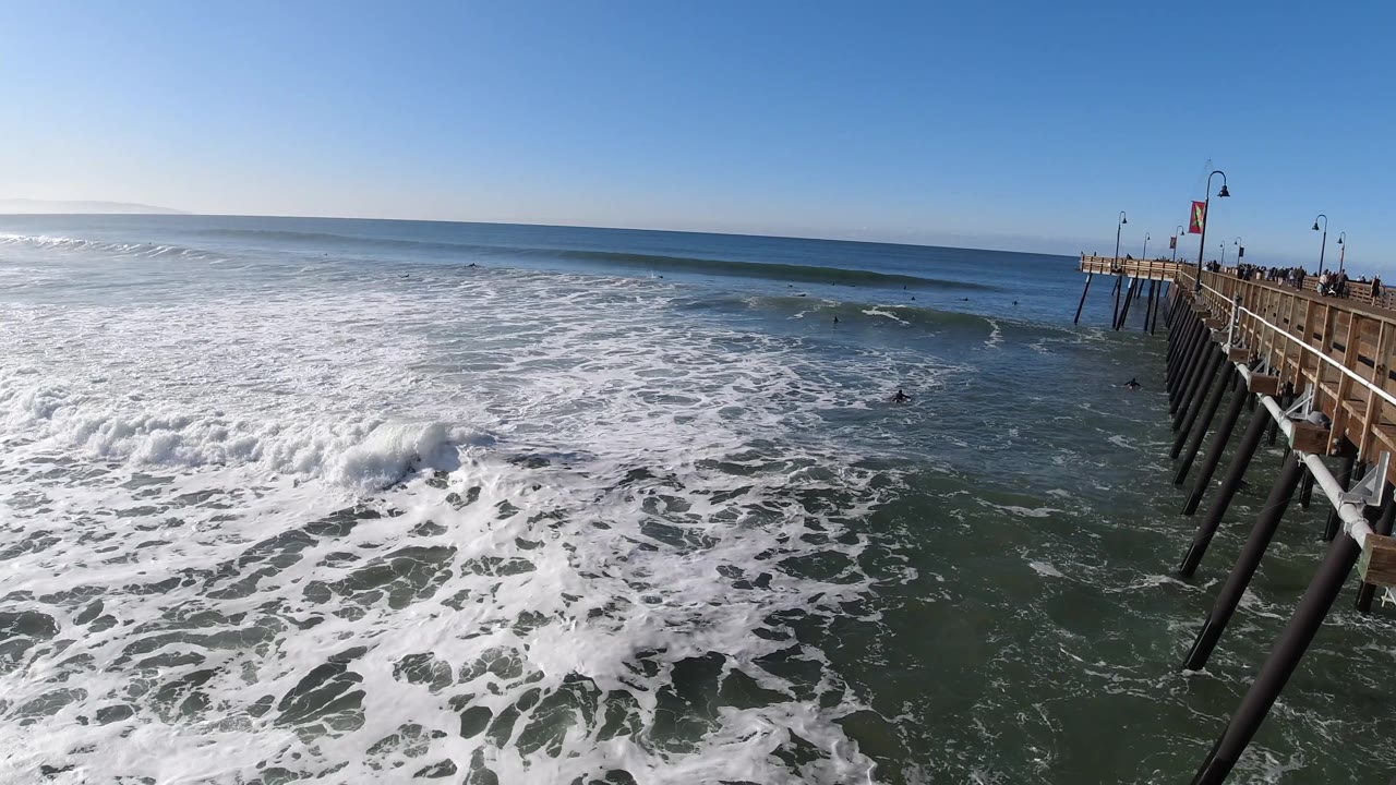 Foamy Pacific water spreads across the foreground at Pismo Beach, with small breaking waves offshore, the pier on the right, and no visible people.