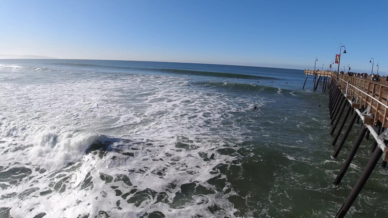 A larger wave breaks near the lower left of the Pismo Beach frame, with churning whitewater across the ocean, the pier to the right, and no visible people.