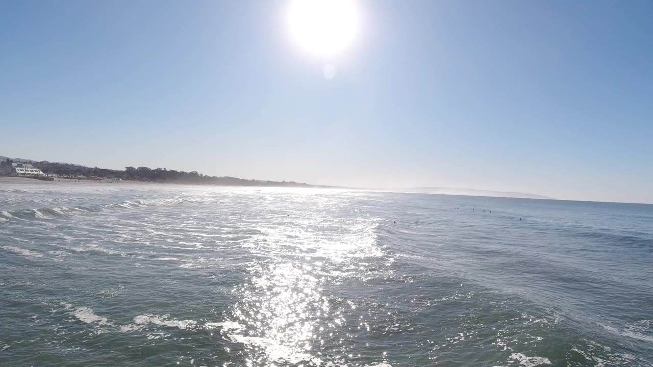 Bright sun reflects across choppy ocean water toward the distant Pismo Beach shoreline, with no clearly visible people.