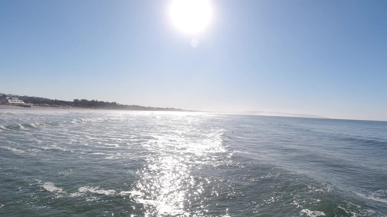 The sun sits high above rippled blue water as a bright reflection leads toward the Pismo Beach coast, with no clearly visible people.