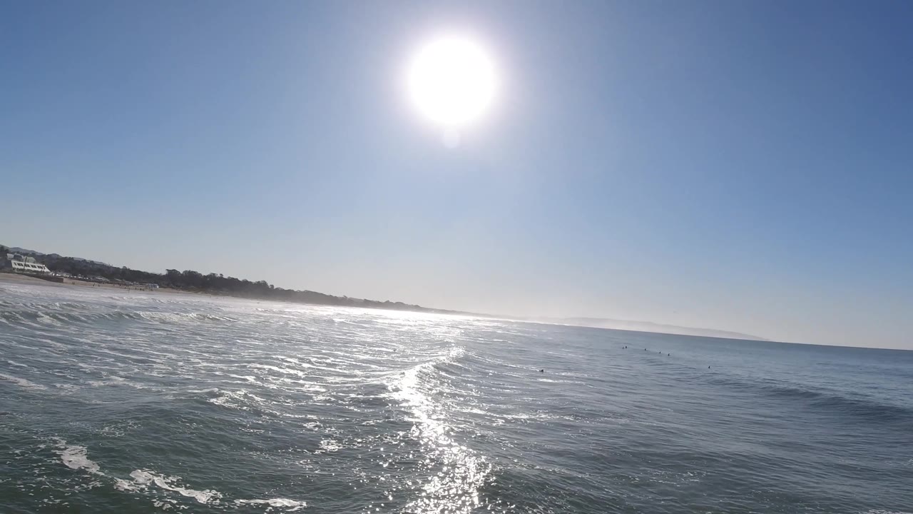 Small rolling waves move under a high white sun, with the Pismo Beach shoreline slanting across the background and no clearly visible people.