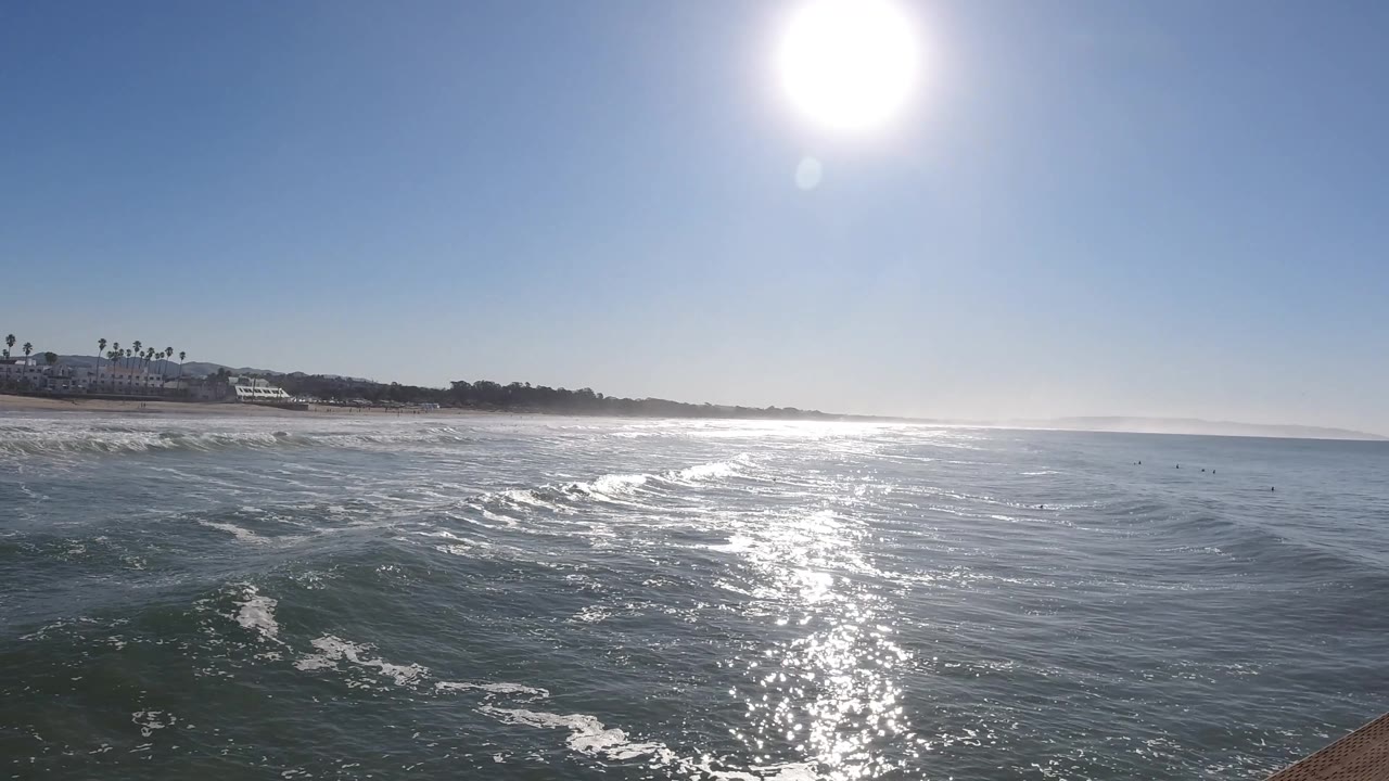 A wider water view at Pismo Beach, California shows the sun high overhead, broken surf lines in the foreground, hazy horizon to the right, distant shore at left, and no visible people.