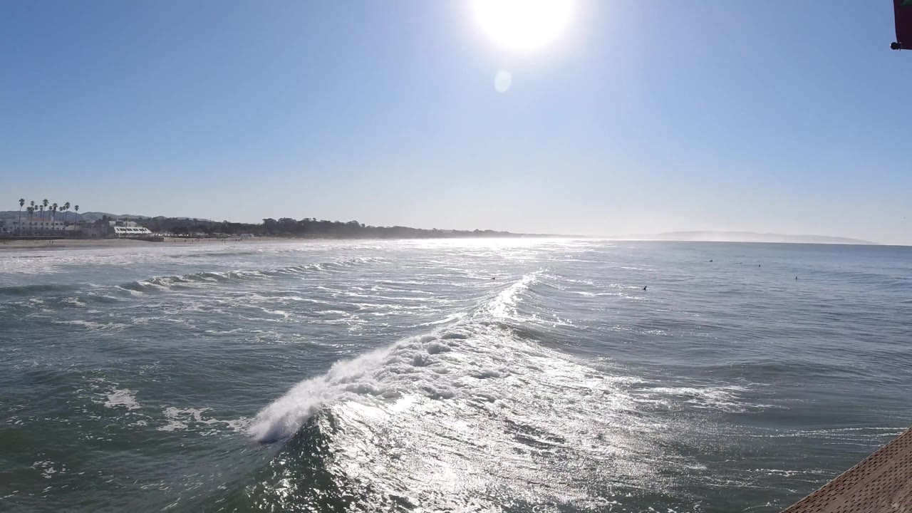 A diagonal band of white surf cuts through reflective water in Pismo Beach, California, with the beach and low buildings left of frame and no visible people.