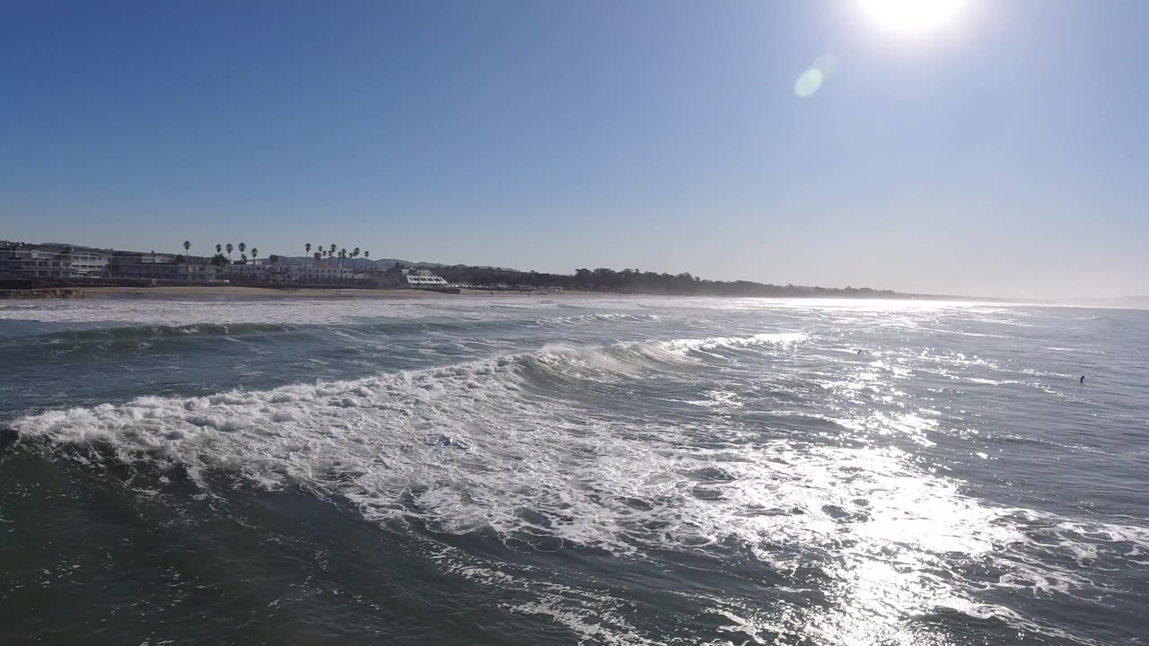 Pismo Beach waves spread across the foreground while sunlight glitters on the water and low coastal buildings sit along the distant left shoreline, with no visible people.