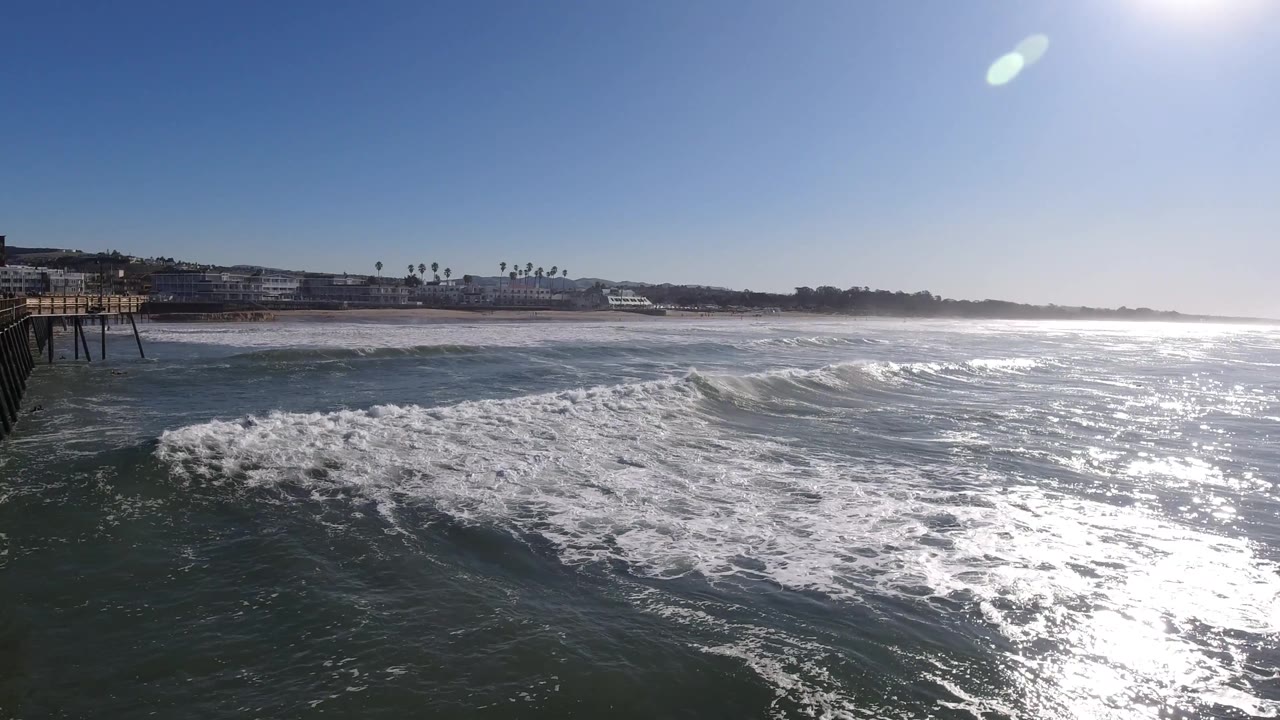A pier or boardwalk structure enters at the left edge over Pismo Beach surf, with breaking waves, distant shoreline buildings, intense sun glare, and no visible people.