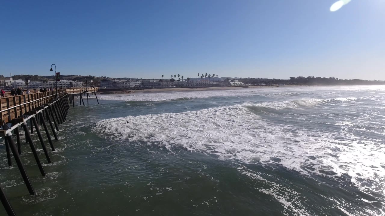 The left side shows a boardwalk or pier with railing and vertical supports above Pismo Beach surf, while whitewater fills the midground and distant buildings line the shore, with no visible people.