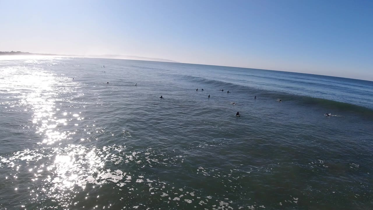 Wide elevated view over Pismo Beach water with bright sun reflection on the left, distant surfers scattered near a small wave, and open sky above the horizon.