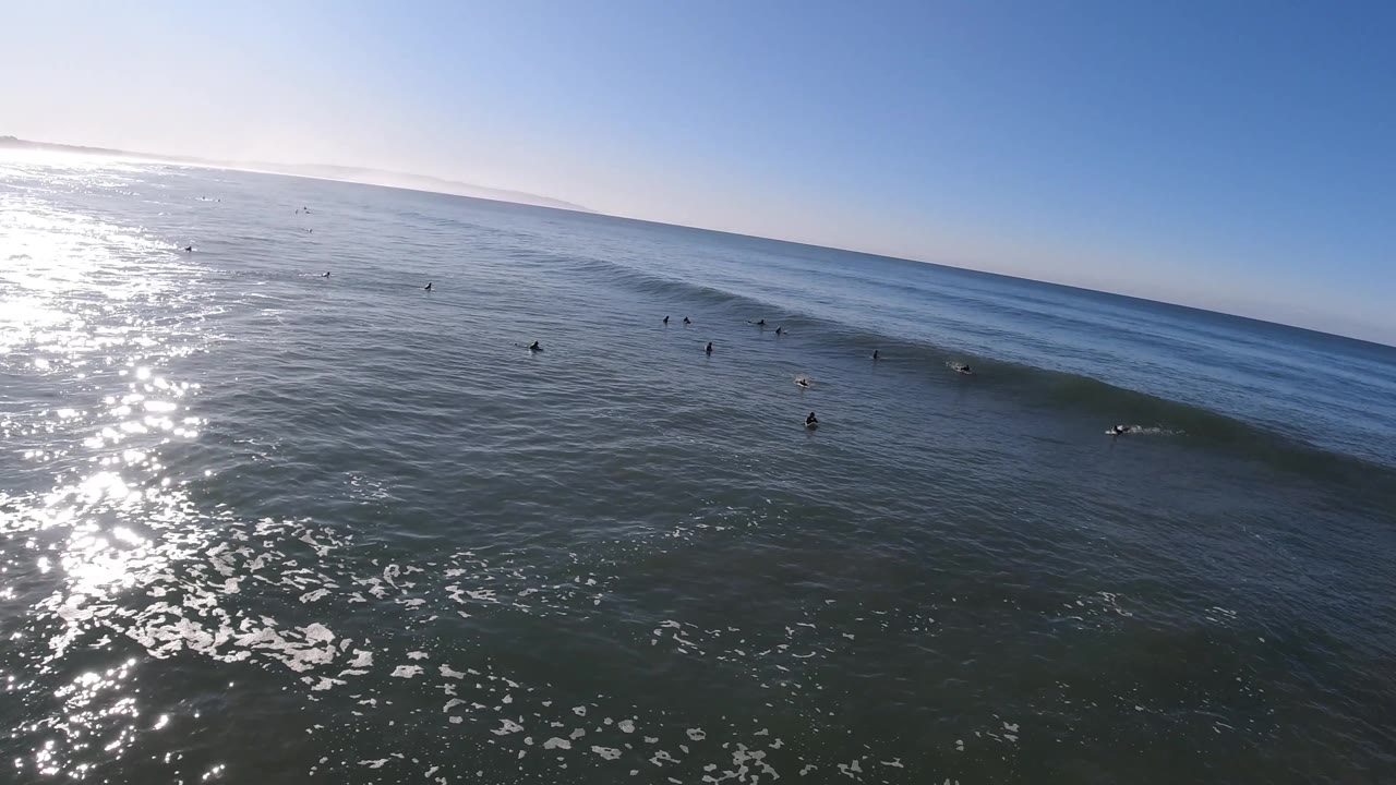 Elevated Pismo Beach view with bright water glare on the left, distant surfers spaced along a wave ridge, and a broad blue sky over the ocean horizon.