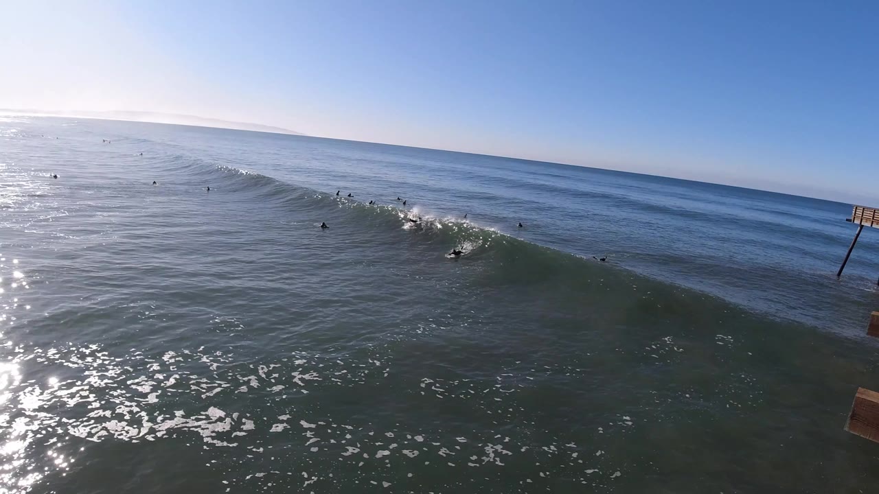 Aerial view over Pismo Beach water with a green wave lifting across the frame, small surfers offshore, bright sun glare on the left, and open blue ocean beyond.
