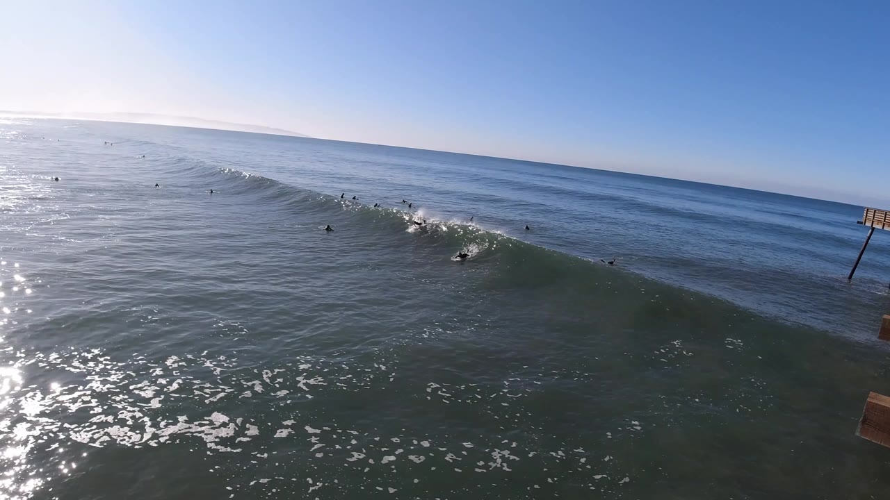 Aerial Pismo Beach surf frame with a surfer low on the wave face, whitewater starting behind them, other surfers farther out, and sun sparkle across the left side of the water.