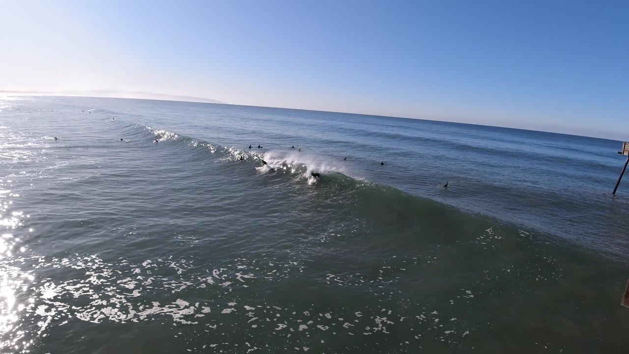 Aerial view in Pismo Beach with the wave crest throwing whitewater across the right side, one surfer on the face, scattered surfers behind the break, and a clear blue sky above the horizon.