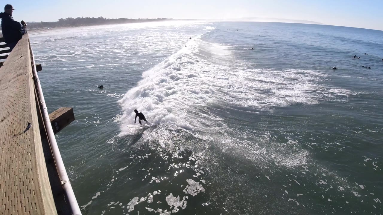 A wooden pier and railing fill the left side as a surfer rides below and a line of whitewater stretches across the ocean at Pismo Beach, California.