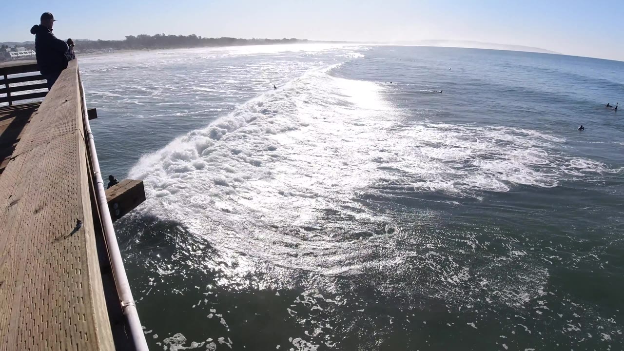 Pier edge and ocean whitewater with surfers offshore.