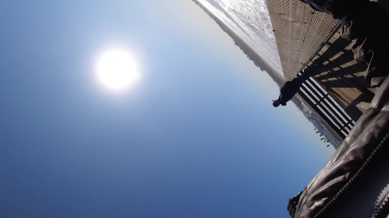 At Pismo Beach, a tilted upward frame catches the bright sun over blue sky, the underside of a wooden pier rail, surf at the edge, and part of a person close to the camera.