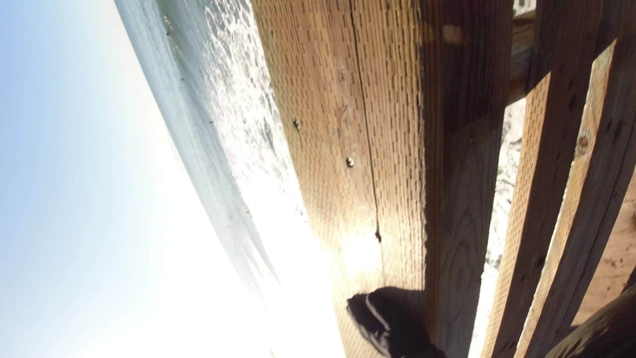 At Pismo Beach, a close oblique angle shows vertical wooden boards and pier railing with harsh sun reflection, a strip of ocean, and part of a person near the lower edge.