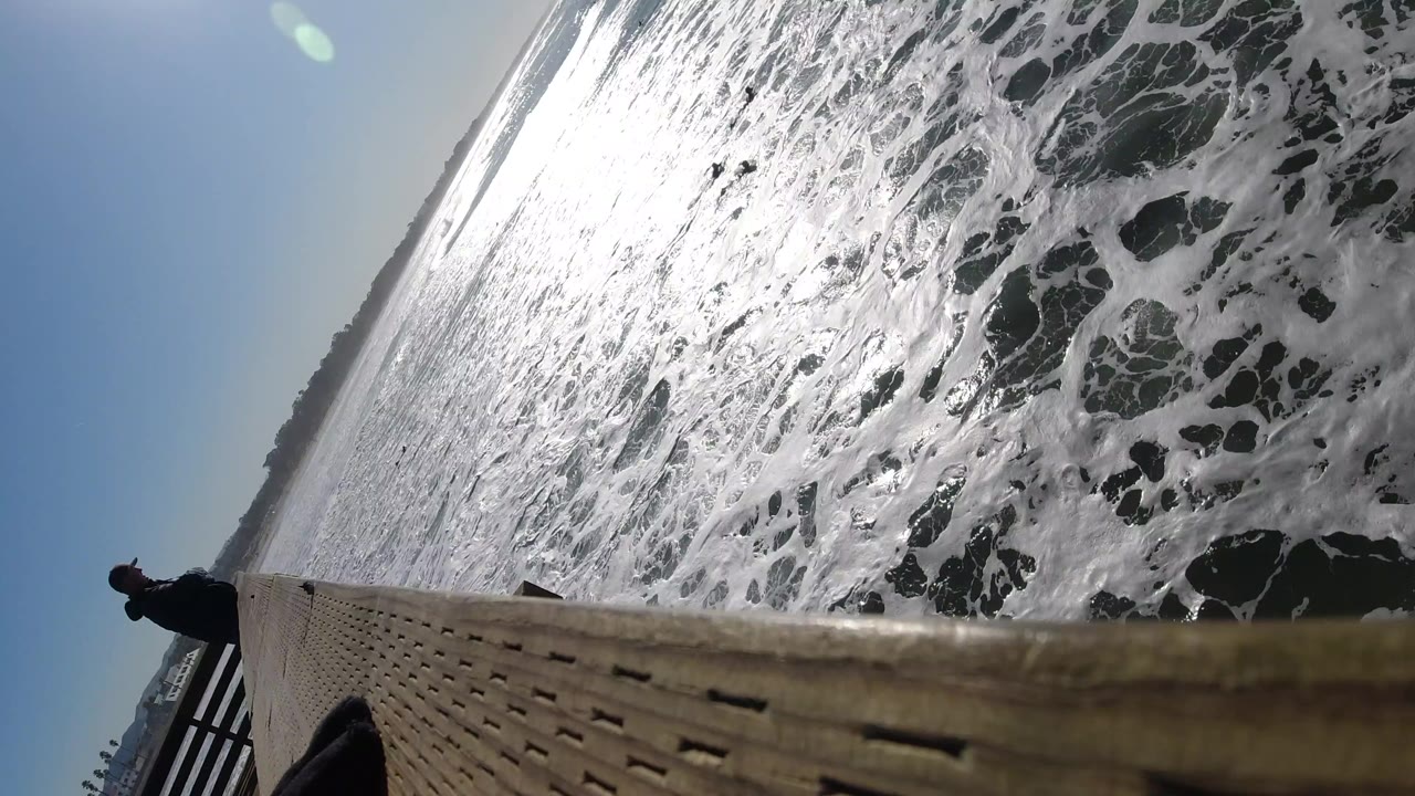 At Pismo Beach, California, foamy surf fills a tilted frame beside a wooden pier edge, with a small person visible near the left railing.