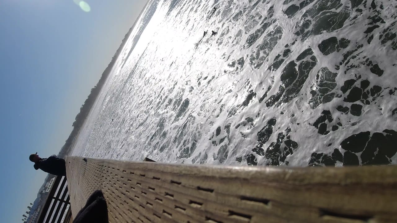 At Pismo Beach, California, sunlight catches dense ocean foam beside the wooden dock edge, with a silhouetted person visible at the far left.