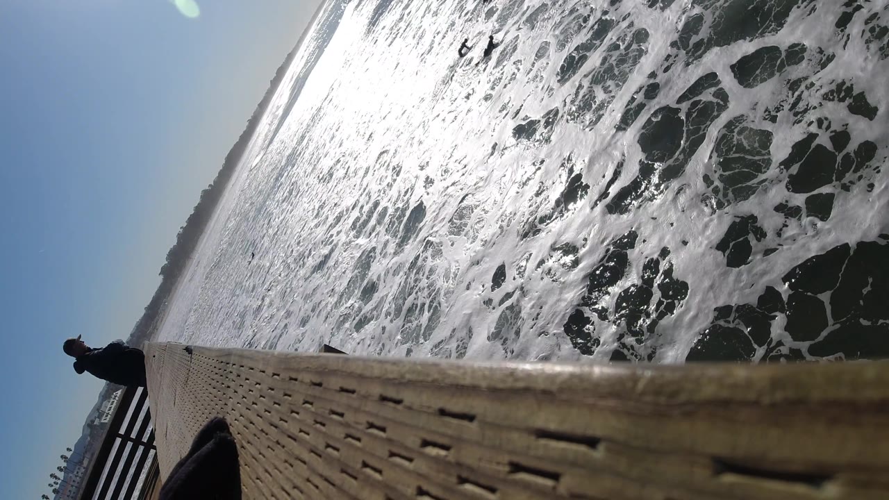 Tilted view of surf foam, blue sky, railing, and wooden pier edge.