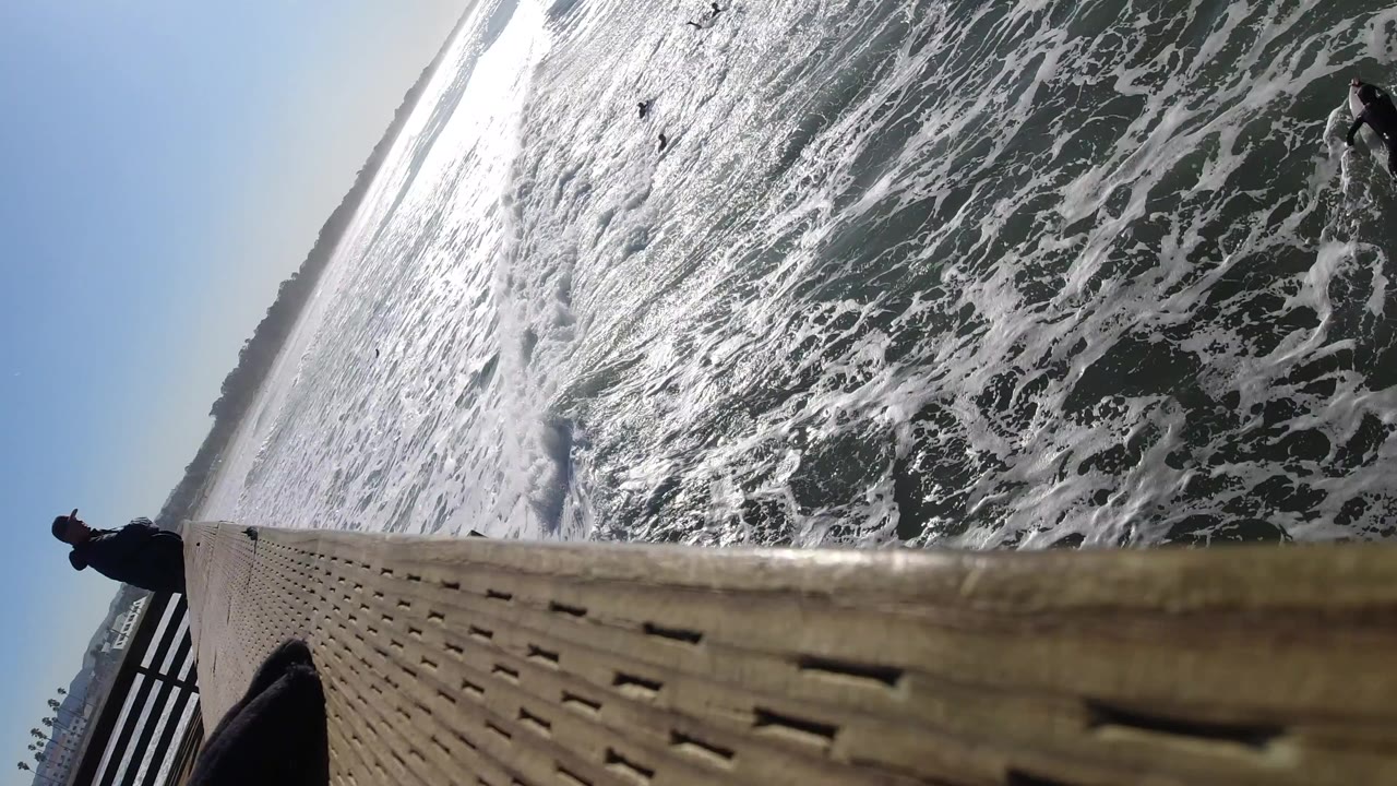 At Pismo Beach, California, the wooden deck fills the lower frame while foamy water and several surfers sit or paddle below under strong sunlight.