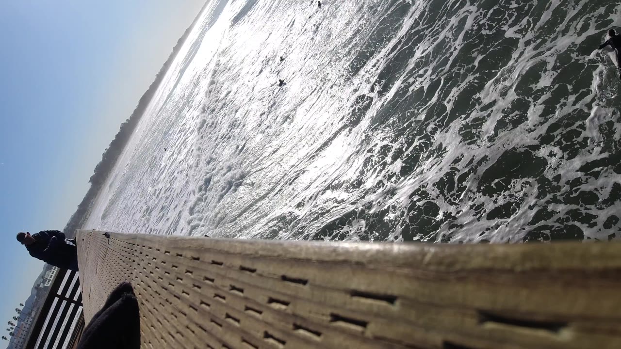 At Pismo Beach, California, a person stands near the left railing while surfers paddle through white foam beside the wooden deck in a steep diagonal composition.