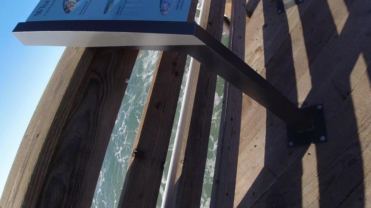 A Pismo Beach boardwalk frame shows vertical railings, deck shadows, a posted sign at the top edge, and ocean visible through the gaps with no visible people.