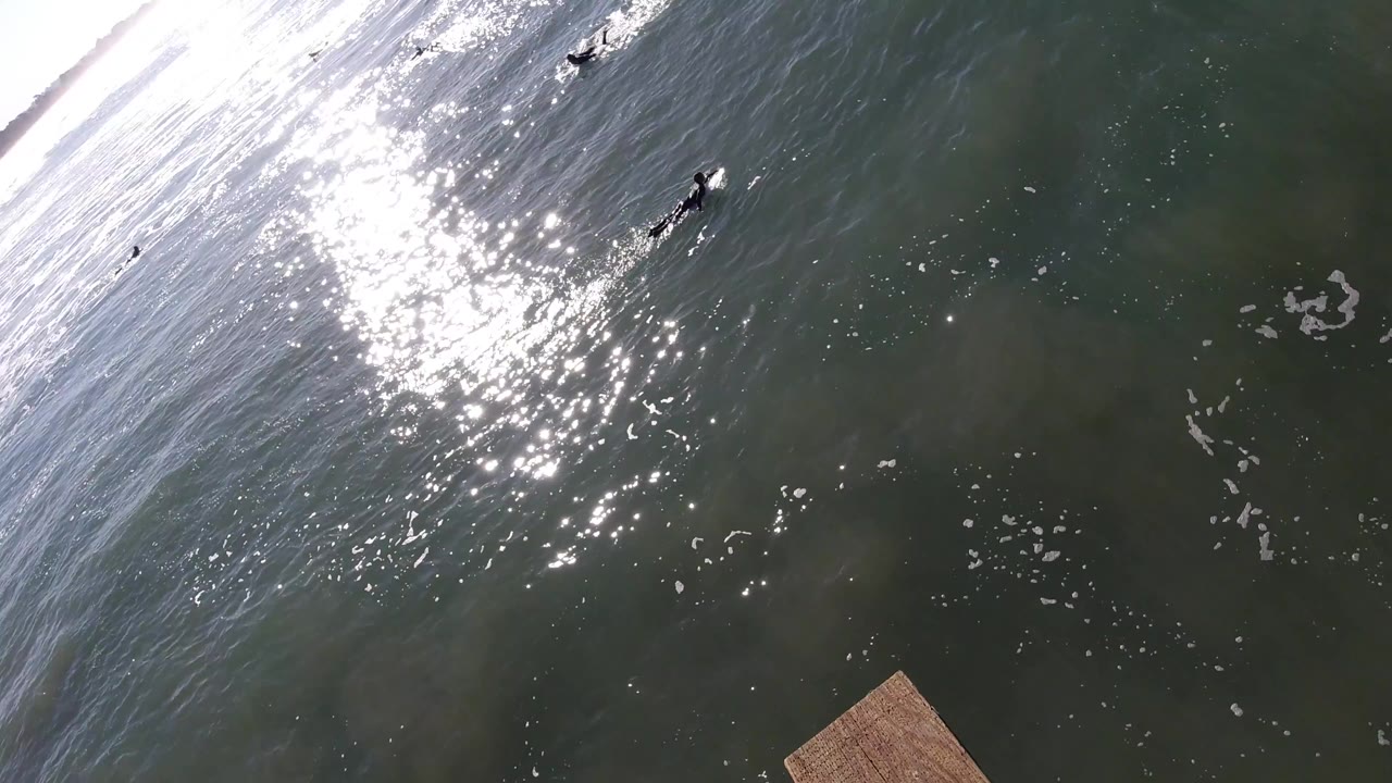High angled view over Pismo Beach ocean water with bright sun glare, a diagonal breaking wave, small surfers near the upper frame, and no visible people close to camera.
